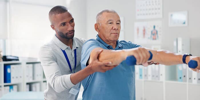 A physical therapist supports an older man’s arm as he performs a shoulder exercise with small dumbbells in a clinic.