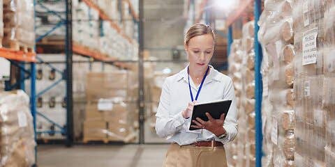 A woman uses a tablet while standing between rows of stacked inventory in a warehouse.