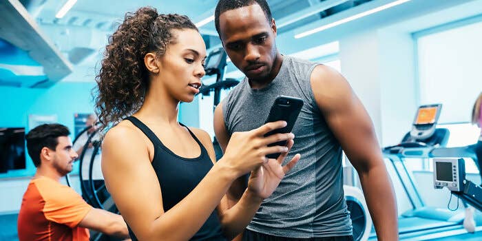 A woman shows a smartphone to a man in workout clothes, with exercise equipment and another gym-goer in the background.